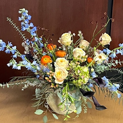 Cream roses blue delphinium and orange ranunculus in a burlap vessel showcasing Cary luxury flowers