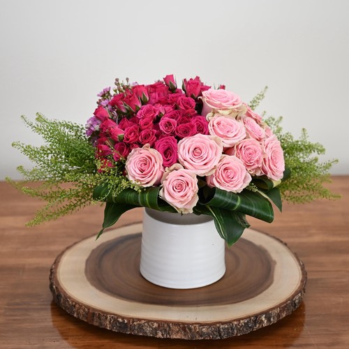 Pink and hot pink roses with fern in a white vessel on a wood slice showcasing Cary luxury flowers