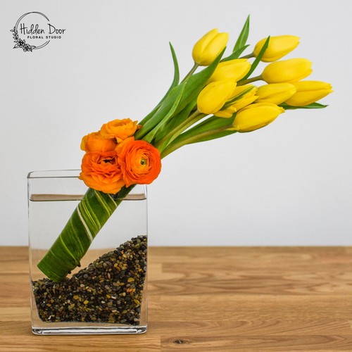 Yellow tulips and orange ranunculus in a modern glass vase with gravel showcasing Cary luxury flowers