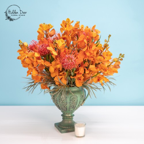 Orange mokara orchids and pincushion protea in a teal vintage urn for luxury flower delivery Cary